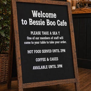 May include: A dark brown wooden A-frame chalkboard sign with white text that reads: Welcome to Bessie Boo Cafe, PLEASE TAKE A SEA T, One of our members of staff will come to your table to take your order, HOT FOOD SERVED UNTIL 2PM, COFFEE & CAKES, AVAILABLE UNTIL 3PM.