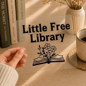 May include: A square transparent acrylic sign featuring the black text Little Free Library and a graphic of an open book with flowers growing from the pages. The sign is held up, displaying the bold lettering and the floral book illustration.