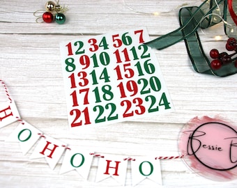 May include: A white card with red and green numbers from 1 to 24. The card is on a white wooden surface with a red and white string banner that says "HO HO HO".