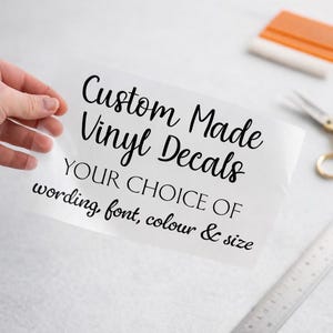 May include: A clear transfer sheet featuring black text that reads Custom Made Vinyl Decals YOUR CHOICE OF wording, font, colour & size. The sheet is held up, displaying the lettering clearly against the transparent backing material.
