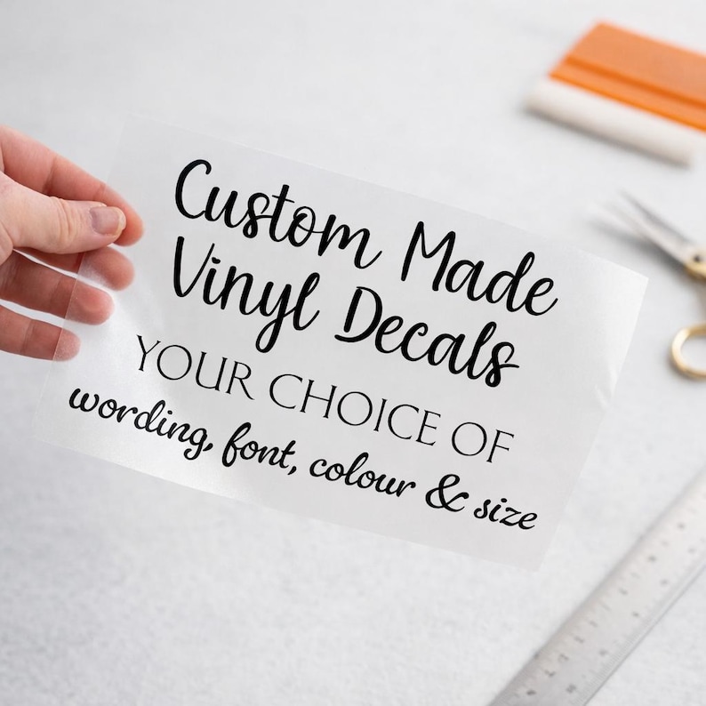 May include: A clear transfer sheet featuring black text that reads Custom Made Vinyl Decals YOUR CHOICE OF wording, font, colour & size. The sheet is held up, displaying the lettering clearly against the transparent backing material.