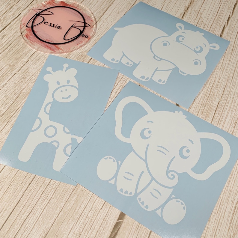 May include: Three light blue vinyl decals featuring white silhouettes of a giraffe, a hippopotamus, and an elephant. A circular clear acrylic disc with the black text Bessie Boo and pink brush strokes is positioned in the upper left corner.