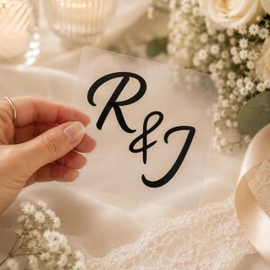 May include: A square, semi-transparent acrylic sign featuring the black printed letters R & J in a flowing script font. The sign is held up, displaying the bold, dark lettering clearly against the frosted, matte surface of the material.