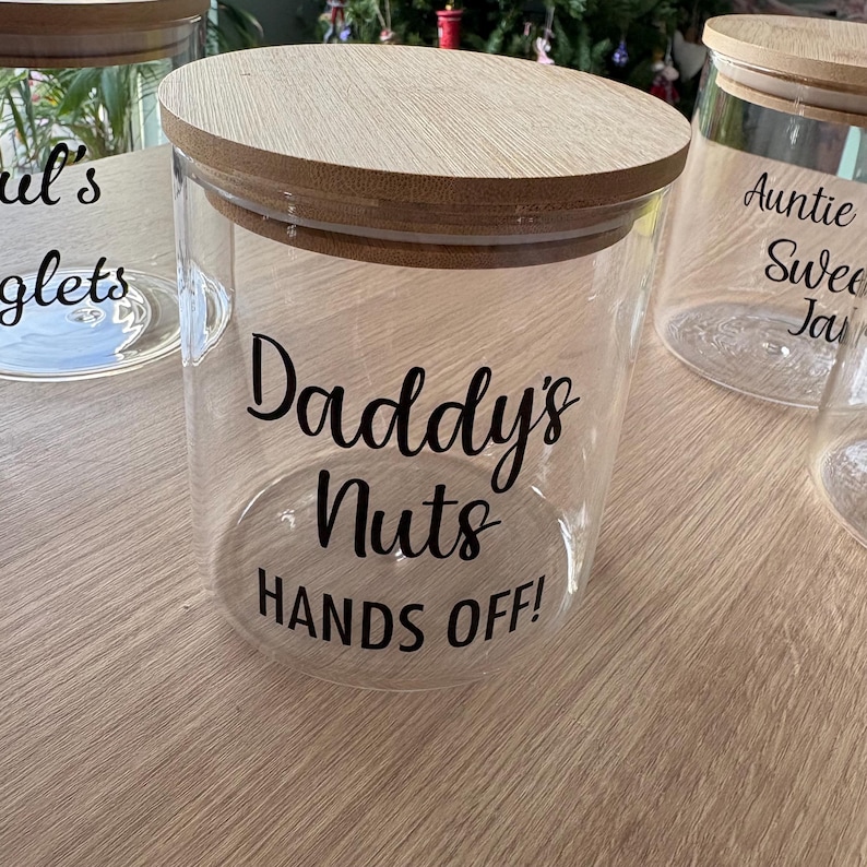 May include: A clear glass storage jar with a round wooden lid. The front of the jar features black lettering that reads Daddy's Nuts HANDS OFF! The jar is cylindrical with a smooth surface and a flat base, designed for holding small items.