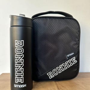 May include: Black water bottle and lunch bag set. The water bottle has a black lid and the name "RONNIE" in white block letters. The lunch bag is black with a honeycomb pattern and the name "RONNIE" in white block letters.