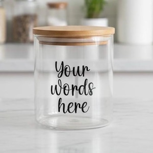 May include: A clear glass jar with a wooden lid. The jar has the words "Your words here" in black script. The jar is cylindrical and appears to be empty. The background is blurred, suggesting a kitchen setting.