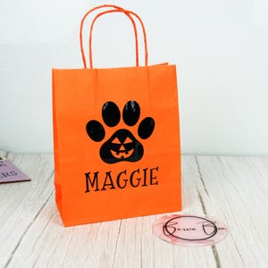 May include: An orange paper gift bag featuring a black paw print design with a jack-o'-lantern face and the name "MAGGIE" printed below. The bag has orange handles and is suitable for Halloween.