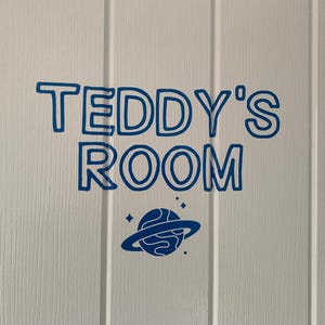 May include: A blue vinyl decal applied to a white paneled door. The decal features the text "TEDDY'S ROOM" in a bold, rounded, outlined font, positioned above a graphic of a planet with rings and small star accents in the same blue color.