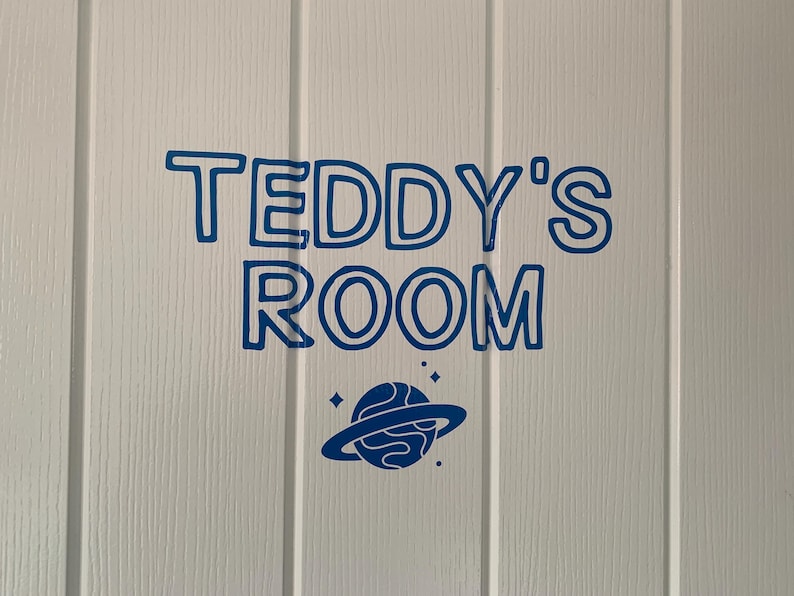 May include: A blue vinyl decal applied to a white paneled door. The decal features the text "TEDDY'S ROOM" in a bold, rounded, outlined font, positioned above a graphic of a planet with rings and small star accents in the same blue color.