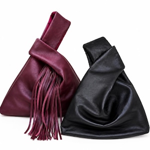 Pode incluir: Duas bolsas de mão estilo nó com silhueta triangular. Uma bolsa é bordô profundo com longas franjas de couro fino penduradas na alça com nó. A segunda bolsa é preta sólida com uma construção de couro liso e dobrado, sem franjas.
