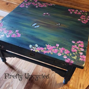 May include: A rectangular wooden coffee table with a dark blue and green painted surface featuring hand-painted pink bougainvillea flowers in the corners and several butterflies scattered across the top. It has a single drawer with a metal ring pull.
