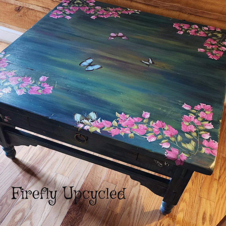 May include: A rectangular wooden coffee table with a dark blue and green painted surface featuring hand-painted pink bougainvillea flowers in the corners and several butterflies scattered across the top. It has a single drawer with a metal ring pull.