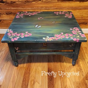 May include: A square wooden coffee table with a dark teal and green painted finish. The top surface features hand-painted pink floral clusters and several butterflies. A single drawer with a circular metal pull is centered on the front apron.
