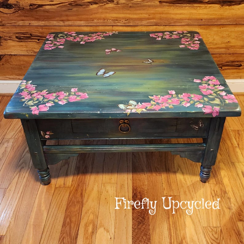 May include: A square wooden coffee table with a dark teal and green painted finish. The top surface features hand-painted pink floral clusters and several butterflies. A single drawer with a circular metal pull is centered on the front apron.