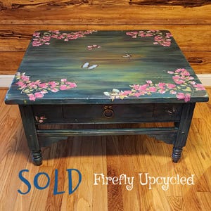 May include: A square wooden coffee table with a dark teal and green distressed finish. The top surface features hand-painted pink floral clusters in the corners and several butterflies. It includes a single drawer with a round metal pull.