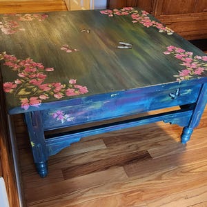 May include: A square wooden coffee table with a dark, multi-toned blue and green painted finish. The surface features hand-painted pink floral clusters in the corners and several butterflies scattered across the top and front apron.