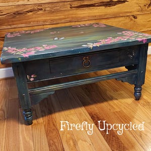 May include: A rectangular wooden coffee table painted in a dark teal finish with hand-painted pink floral clusters and butterflies on the top surface. The table features a single central drawer with a round brass pull and turned legs.