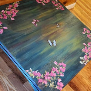 May include: A square wooden table with a dark teal and green brushed finish. The surface is decorated with hand-painted pink bougainvillea flowers along the edges and several butterflies in shades of blue, purple, and white scattered across the center.