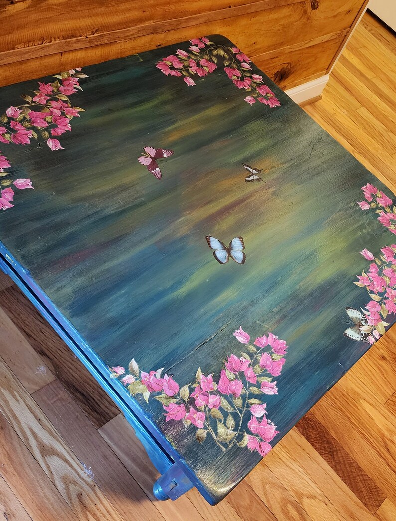 May include: A square wooden table with a dark teal and green brushed finish. The surface is decorated with hand-painted pink bougainvillea flowers along the edges and several butterflies in shades of blue, purple, and white scattered across the center.