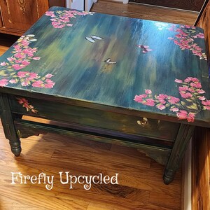 May include: A square wooden coffee table with a dark green and blue brushed finish. The top surface features hand-painted pink floral clusters in the corners and several butterflies scattered across the center. The text Firefly Upcycled is visible.