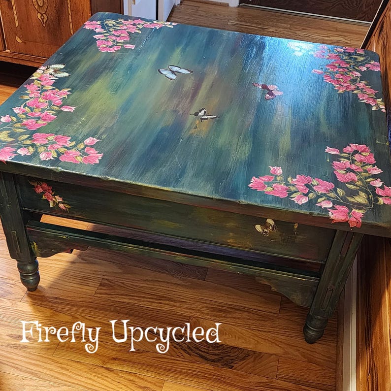 May include: A square wooden coffee table with a dark green and blue brushed finish. The top surface features hand-painted pink floral clusters in the corners and several butterflies scattered across the center. The text Firefly Upcycled is visible.