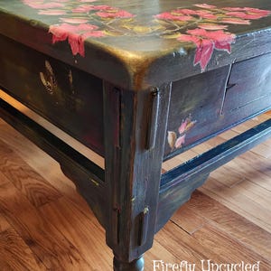May include: A dark-painted wooden coffee table featuring hand-painted pink floral motifs and butterfly illustrations on the top surface and side panels. The table has a rectangular top, sturdy legs, and the text Firefly Upcycled in white.