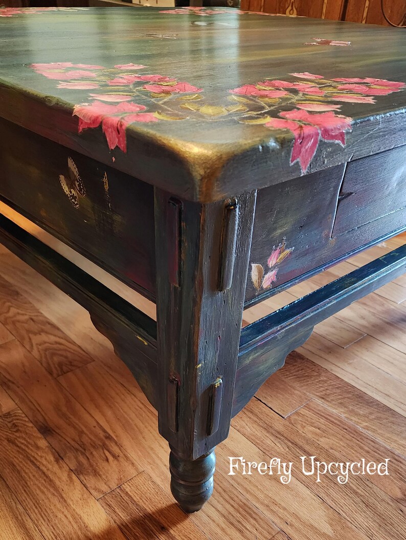 May include: A dark-painted wooden coffee table featuring hand-painted pink floral motifs and butterfly illustrations on the top surface and side panels. The table has a rectangular top, sturdy legs, and the text Firefly Upcycled in white.