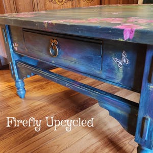 May include: A rectangular wooden coffee table painted in shades of blue and purple with pink floral accents and butterfly motifs. The table features a single drawer with a circular metal ring pull handle and the text Firefly Upcycled.