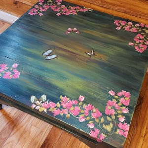May include: A square wooden table with a dark green and blue painted surface featuring hand-painted pink bougainvillea flowers in the corners and several butterflies scattered across the center of the tabletop in a naturalistic arrangement.