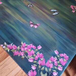 May include: A rectangular table with a dark teal and green brushed finish, featuring hand-painted pink bougainvillea flowers along the edges and three butterflies in flight across the surface. The text Firefly Upcycled is visible.