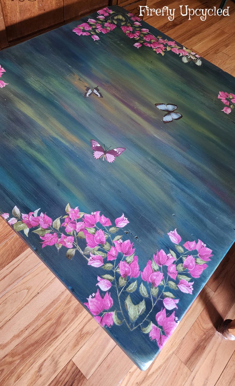 May include: A rectangular table with a dark teal and green brushed finish, featuring hand-painted pink bougainvillea flowers along the edges and three butterflies in flight across the surface. The text Firefly Upcycled is visible.