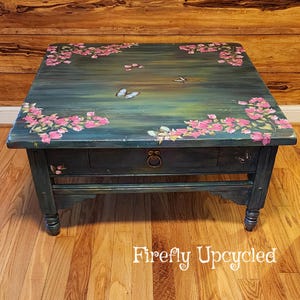 May include: A square coffee table with a dark teal and green painted finish, featuring hand-painted pink bougainvillea flowers in the corners and several butterflies across the surface. The table includes a single drawer with a round metal pull.