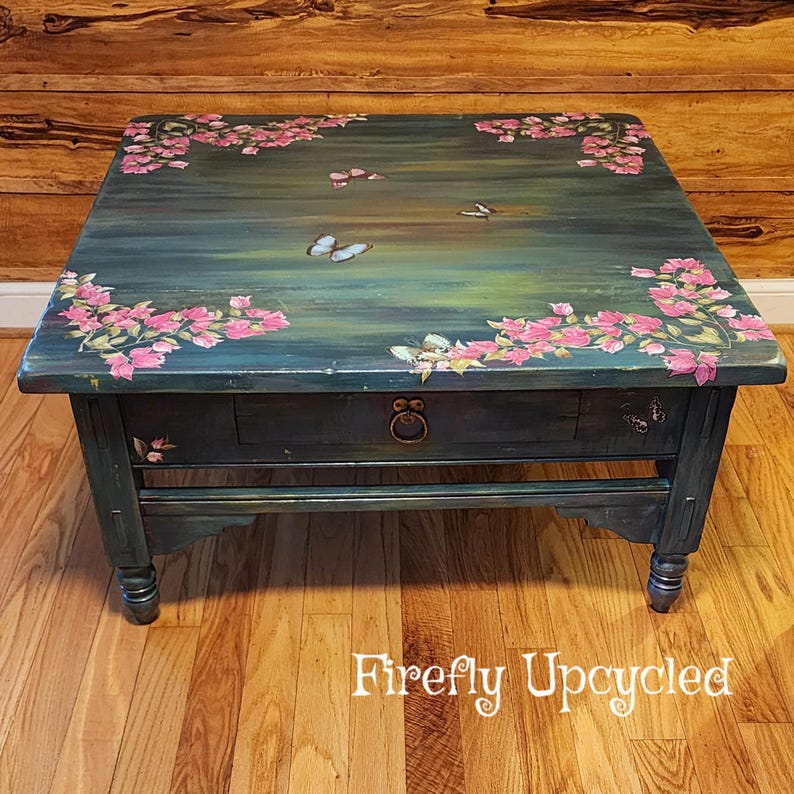 May include: A square coffee table with a dark teal and green painted finish, featuring hand-painted pink bougainvillea flowers in the corners and several butterflies across the surface. The table includes a single drawer with a round metal pull.