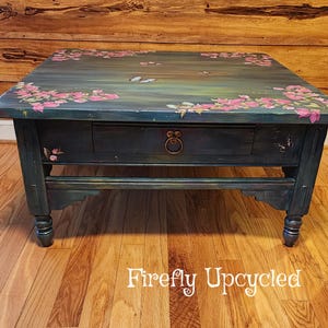 May include: A rectangular wooden coffee table painted in a dark teal finish with hand-painted pink floral clusters and butterflies on the top surface. The table features a single central drawer with a circular metal pull and four turned legs.