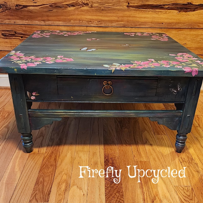 May include: A rectangular wooden coffee table painted in a dark teal finish with hand-painted pink floral clusters and butterflies on the top surface. The table features a single central drawer with a circular metal pull and four turned legs.