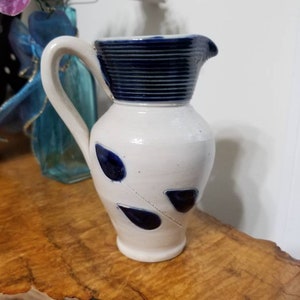 May include: A white ceramic pitcher with a dark blue rim and handle. The pitcher has a series of dark blue teardrop-shaped designs. The pitcher is sitting on a wooden surface.