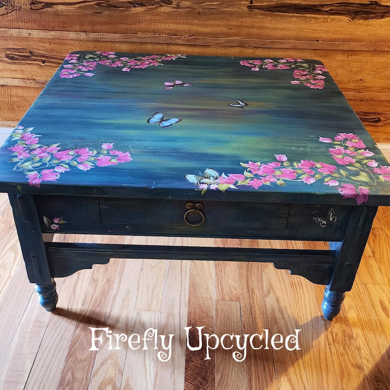 May include: A square wooden coffee table with a dark teal and blue painted finish. The top surface features hand-painted pink floral clusters in the corners and several butterflies. A single drawer with a round metal pull is centered on the front.