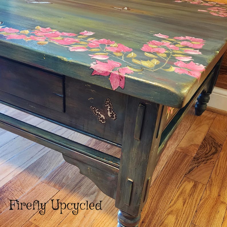 May include: A dark green wooden coffee table featuring hand-painted pink bougainvillea flowers across the top surface and a small butterfly illustration on the front drawer panel. The text Firefly Upcycled is printed in black on the lower left.