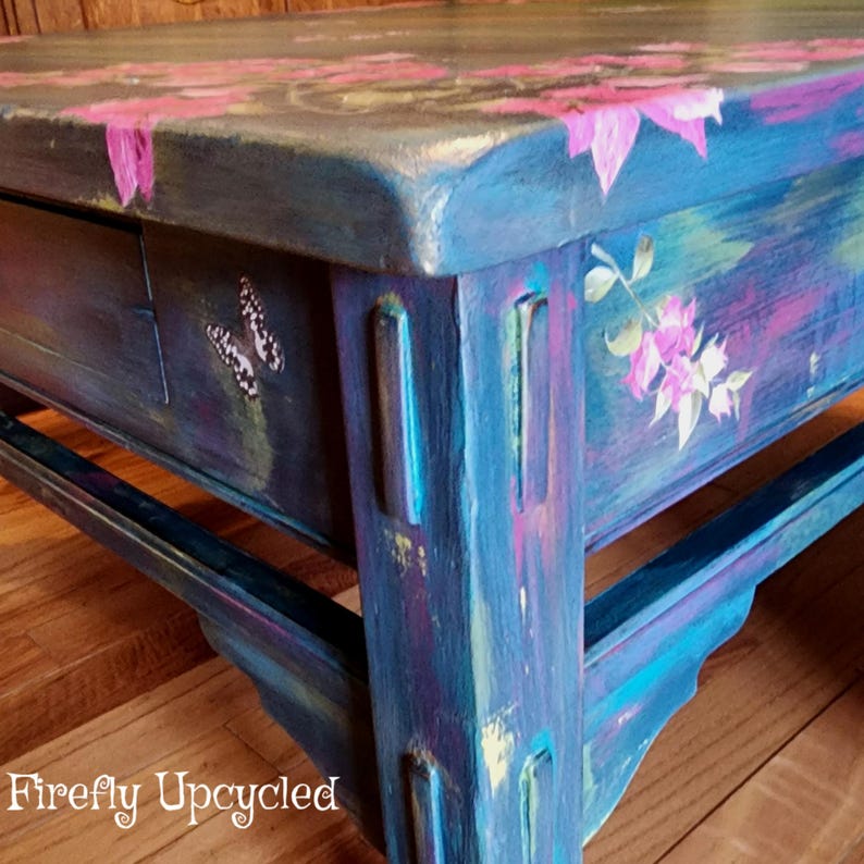May include: A rectangular wooden coffee table painted in shades of blue, purple, and teal with hand-painted pink floral motifs and a butterfly on the drawer front. The table features a textured finish and the text Firefly Upcycled in white.