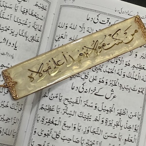 Può includere: Un segnalibro dorato con un nappa e calligrafia araba che recita "Subhana Allah Al-A'zam" su sfondo bianco. Il segnalibro è appoggiato su un libro aperto con testo arabo.