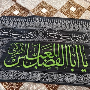 May include: Black textile with Arabic calligraphy in white and green. The central text is in vibrant green, while the borders and other inscriptions are in white. The background is a patterned fabric.