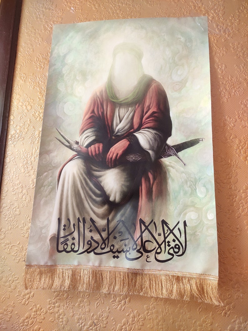 May include: A religious image depicting a figure in a white robe and a green turban, holding a sword. The background is a light blue with white clouds. The image is framed with a gold border and has a fringe at the bottom. The text in the image reads "الْقَوِيُّ عَلَىٰ شَيْئٍ وَهُوَ عَلَىٰ كُلِّ شَيْءٍ قَدِيرٌ".