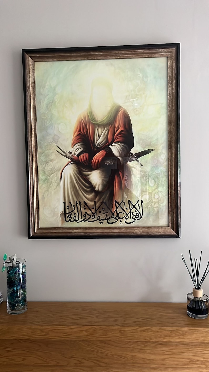 May include: A framed painting depicting a religious figure wearing a red robe, seated with a sword in their lap. The painting is set against a cloudy background with Arabic text at the bottom.