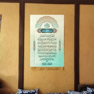 May include: A framed Islamic calligraphy print with a blue and green color scheme. The print features Arabic text and a decorative border. The print is hanging on a yellow wall with a brown wooden frame.