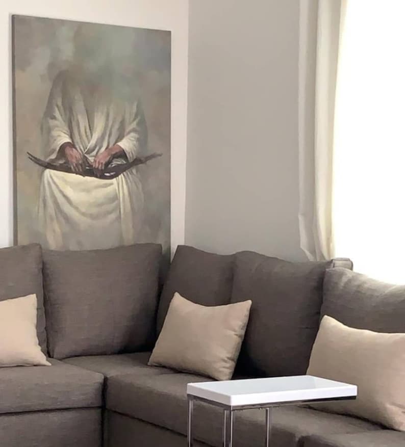 May include: A brown sectional sofa with three beige throw pillows. A white side table with a silver metal frame sits in front of the sofa. A painting of a person in a white robe hangs on the wall behind the sofa.