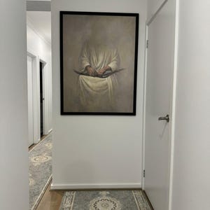 May include: A hallway with white walls and a framed painting of a person holding a book. The hallway has a patterned rug on the floor.