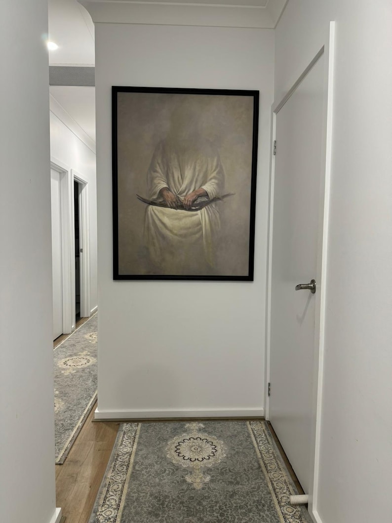 May include: A hallway with white walls and a framed painting of a person holding a book. The hallway has a patterned rug on the floor.