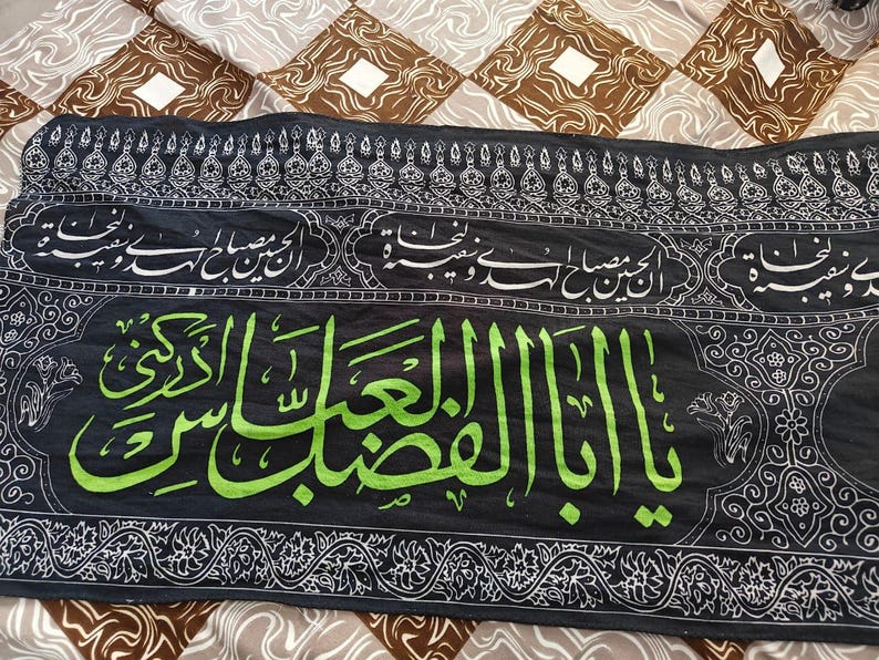 May include: Black textile with Arabic calligraphy in white and green. The central text is in vibrant green, while the borders and other inscriptions are in white. The background is a patterned fabric.