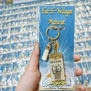 Può includere: Un portachiavi dorato con un fiocco bianco e un ciondolo in resina trasparente con glitter dorati. Il ciondolo presenta calligrafia araba e il testo "Eid al Ghadir". Il portachiavi è confezionato su una carta blu con il testo "Eid-E-Wilayat Mubarak".