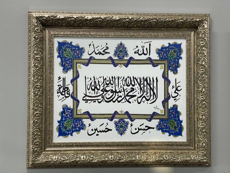 May include: A framed artwork with a white background and intricate blue and gold floral designs. The artwork features Arabic calligraphy in black ink, including the words "Allah" and "Muhammad".